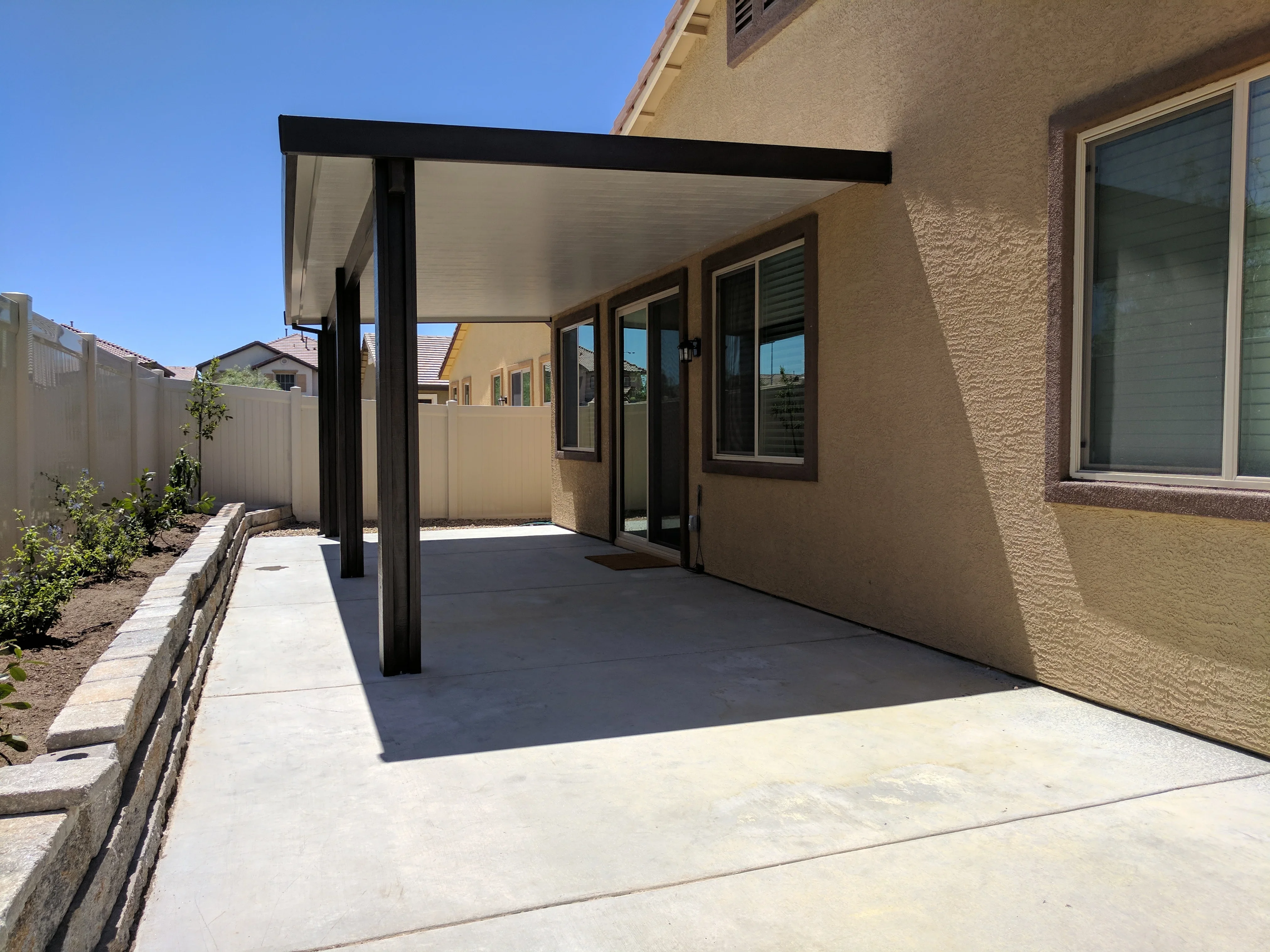 Black pergola with white roof panels over concrete patio adjacent to tan stucco home