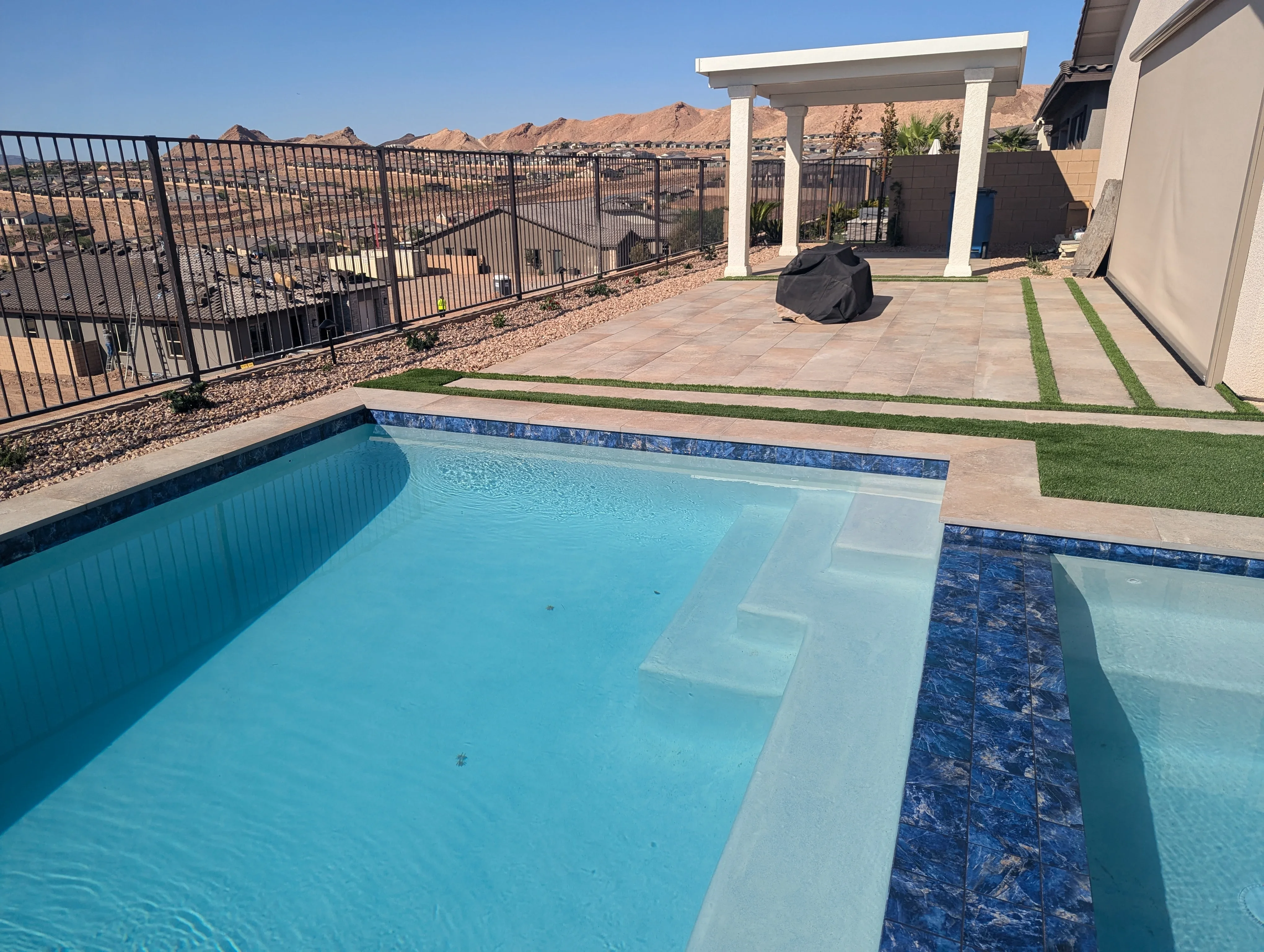 Newly constructed pool with blue tile, concrete patio, white pergola, and black railing on hillside