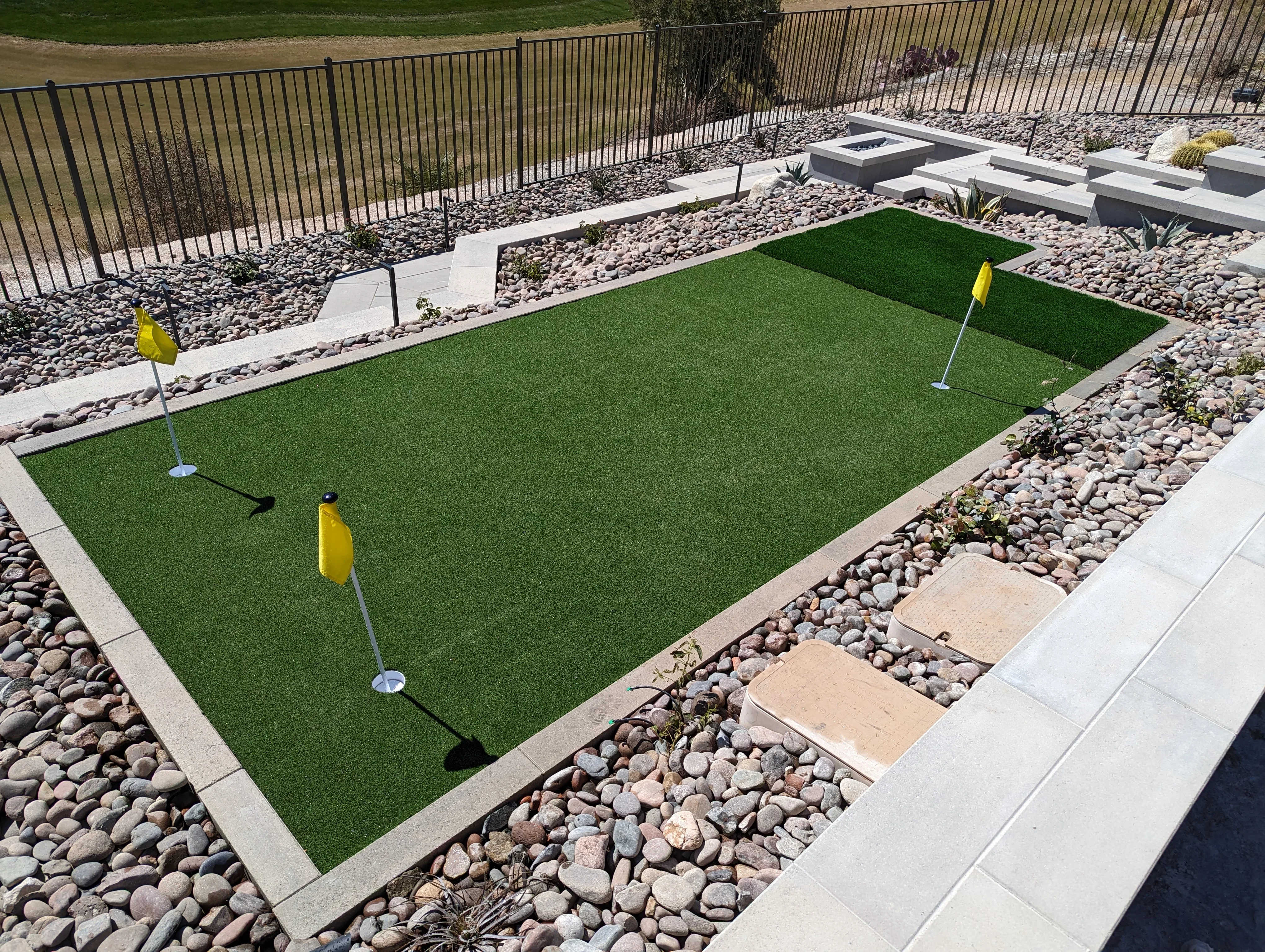 Synthetic putting green with concrete borders, river rock landscaping, and metal flagsticks in backyard setting