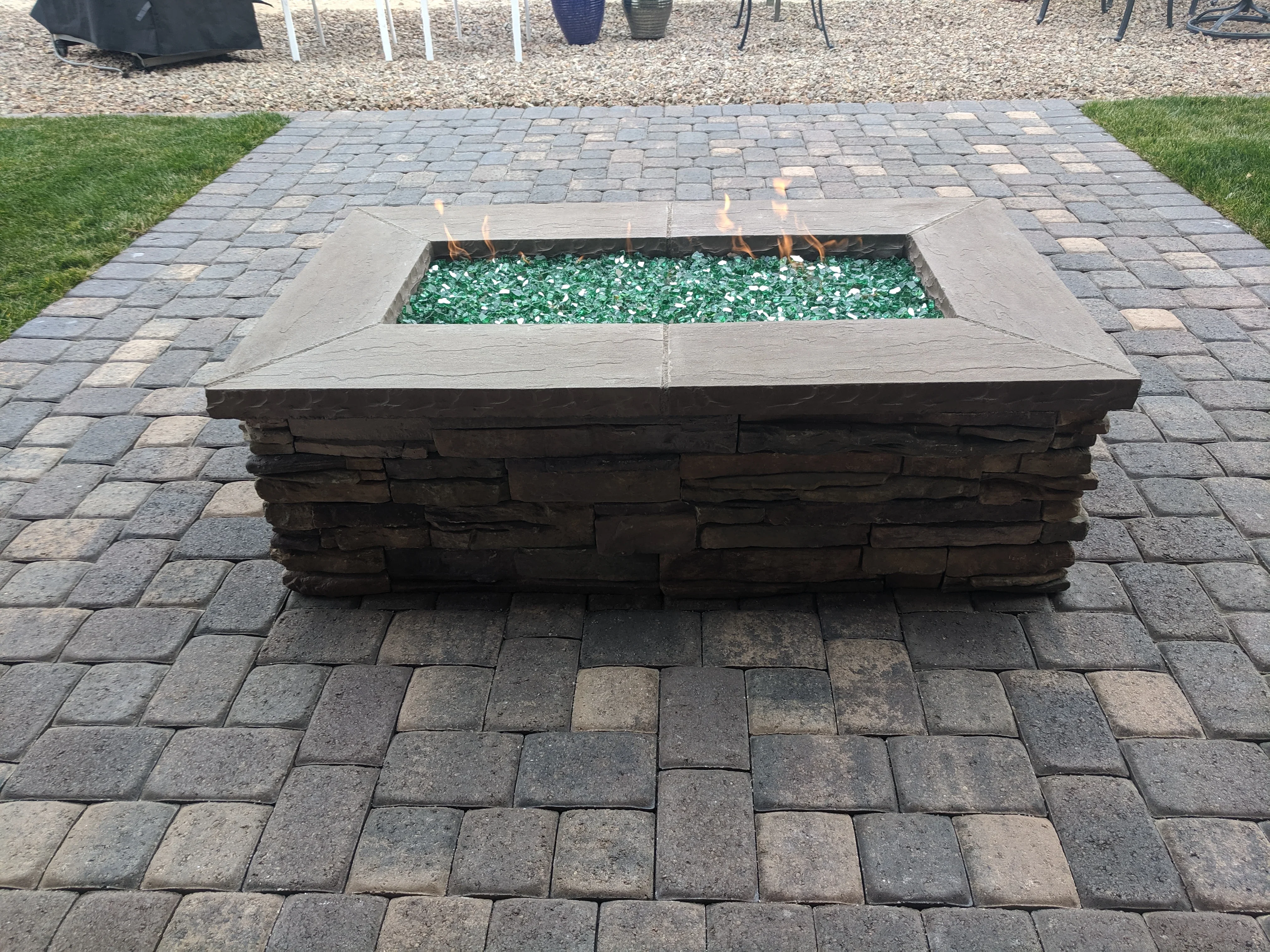 Stone fire pit with turquoise glass media on gray paver patio with lawn borders