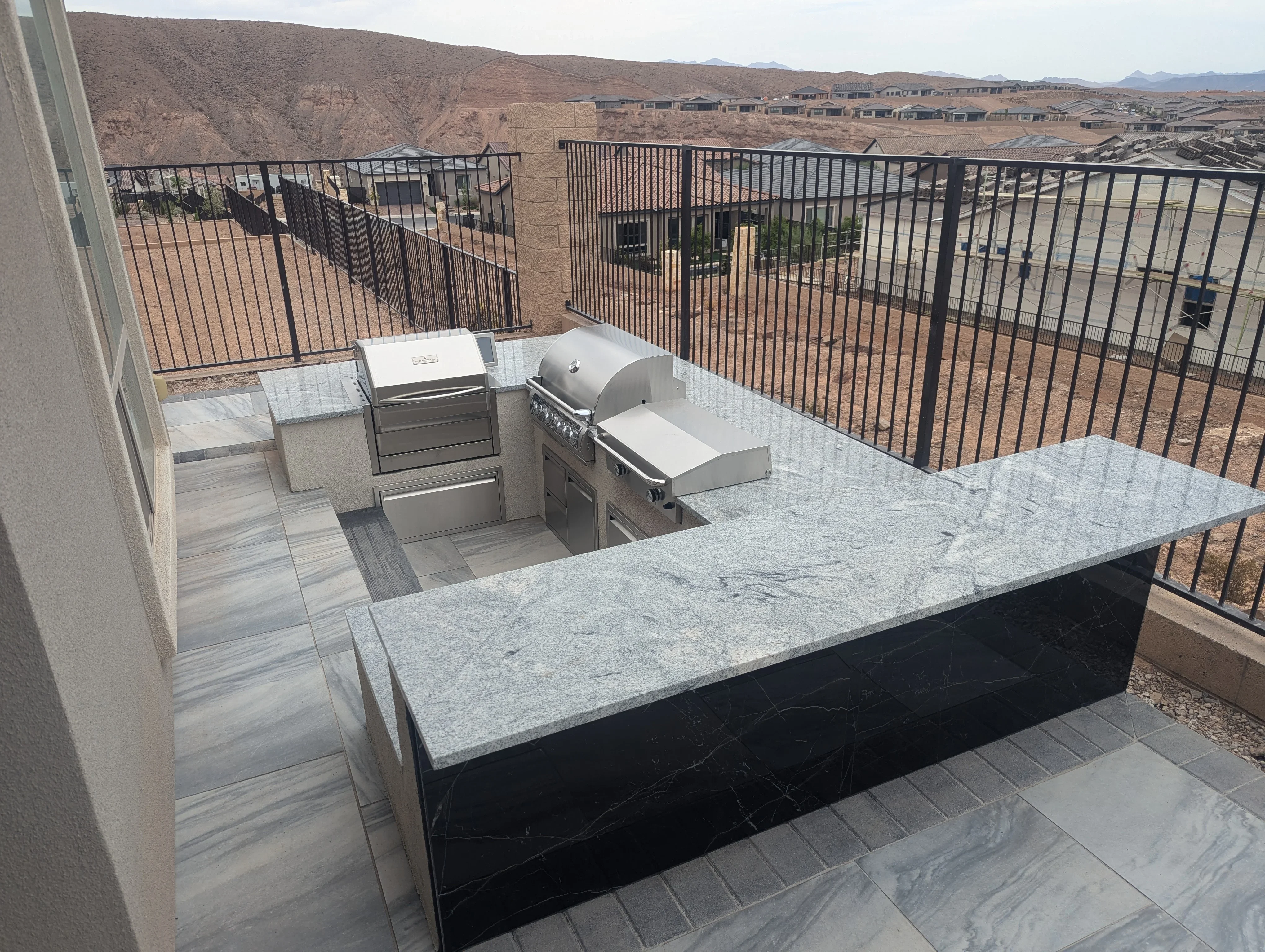 Elevated outdoor kitchen with granite counters, built-in grill, and black metal railings overlooking desert landscape