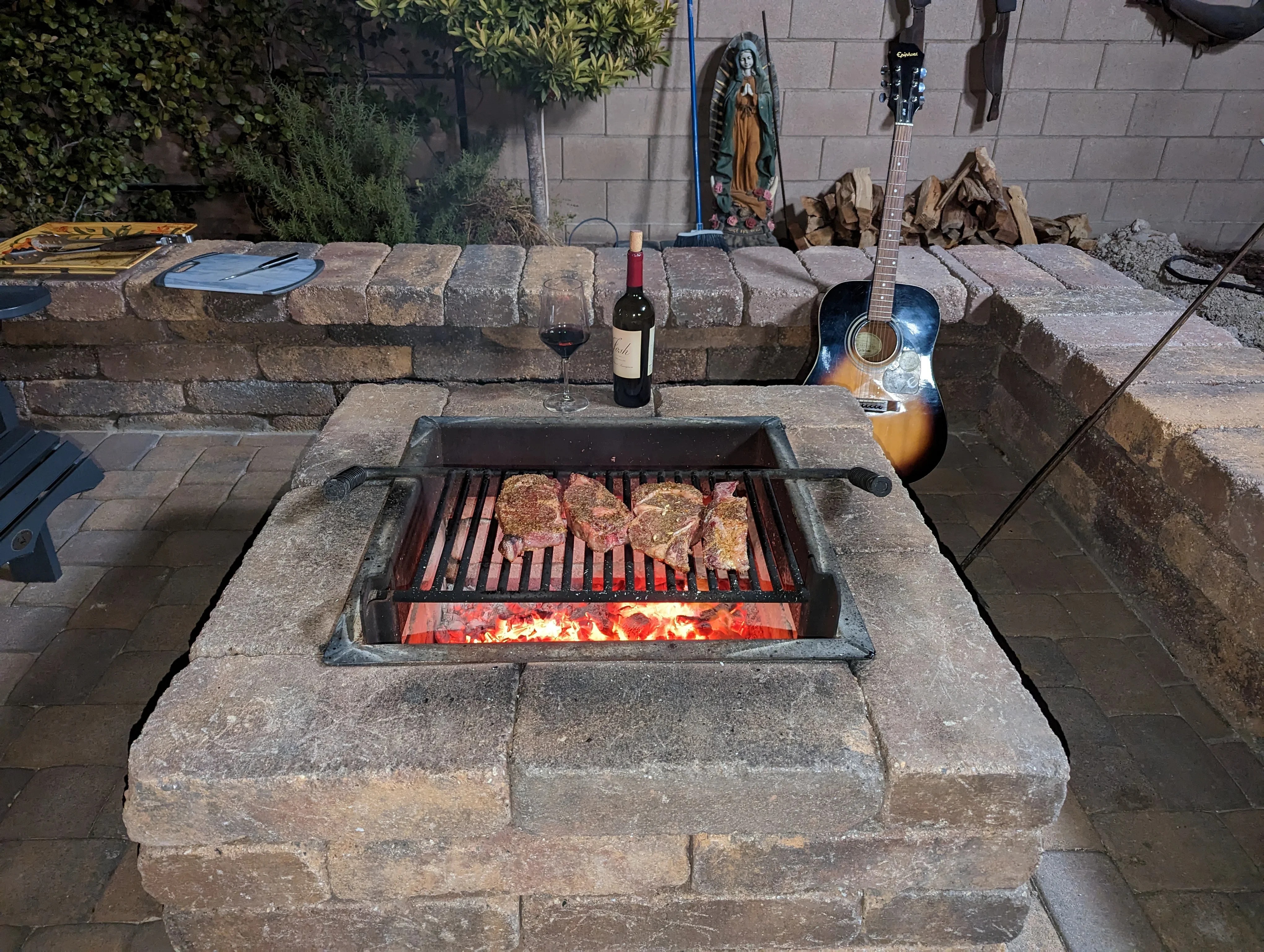 Stone fire pit with metal grill grate, active flames, and surrounding seating wall in backyard patio