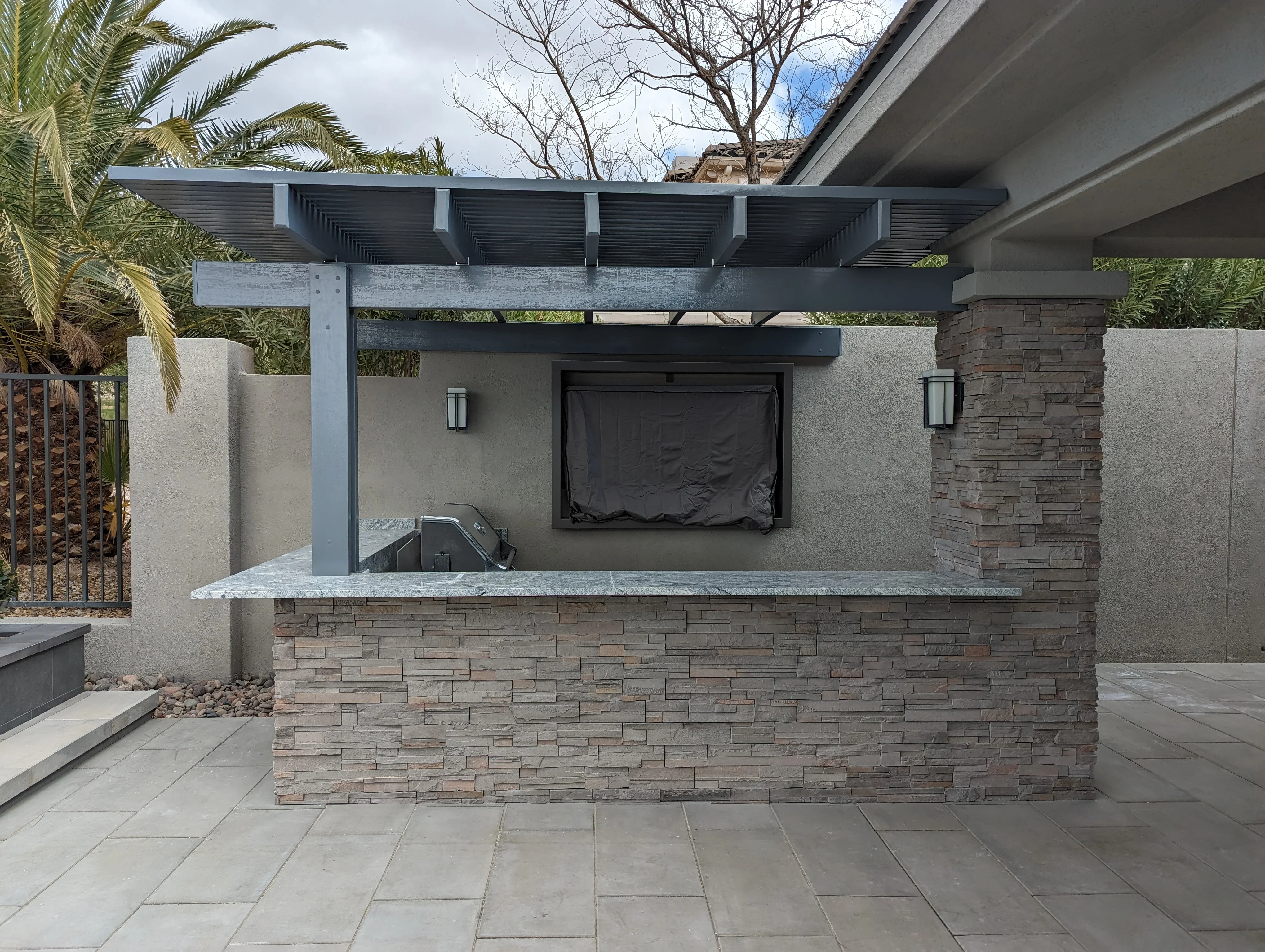 Modern outdoor kitchen with stone base, granite counter, and louvered pergola on paved patio